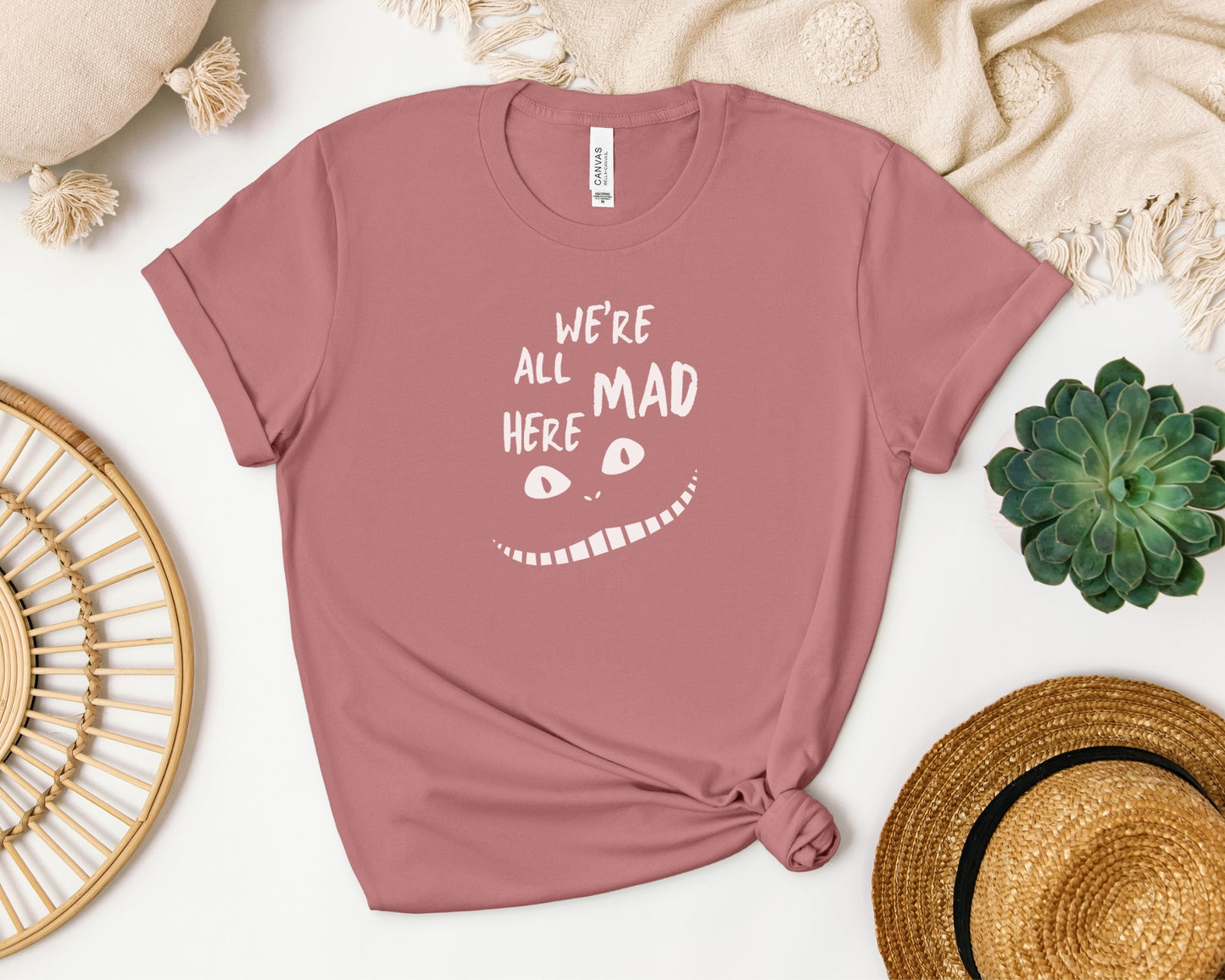 bella and canvas 3001 t-shirt laid flat in mauve with 'we're all mad here' cheshire cat inspired print on chest