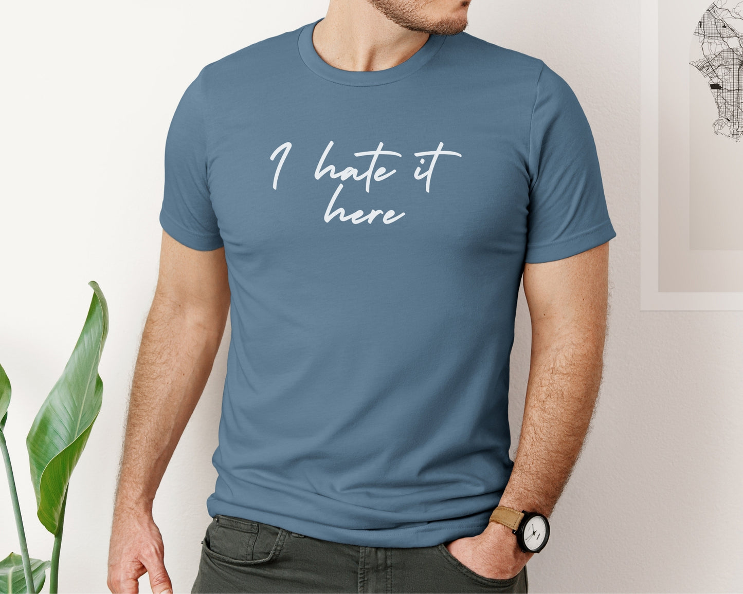 Male model wearing Bella and Canvas 3001 t-shirt in Steel Blue, with TTPD lyric print on chest, T-shirt has 'I hate it here' printed on chest in script style font inspired by Taylor Swift